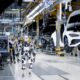 Apptronik and Mercedes-Benz - A humanoid robot walking through a Mercedes-Benz car assembly line in a manufacturing plant.