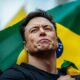 Elon Musk standing in front of a Brazilian flag, looking upwards with a serious expression, possibly pondering the recent feud with Elon Musk and how Brazil blocks X.