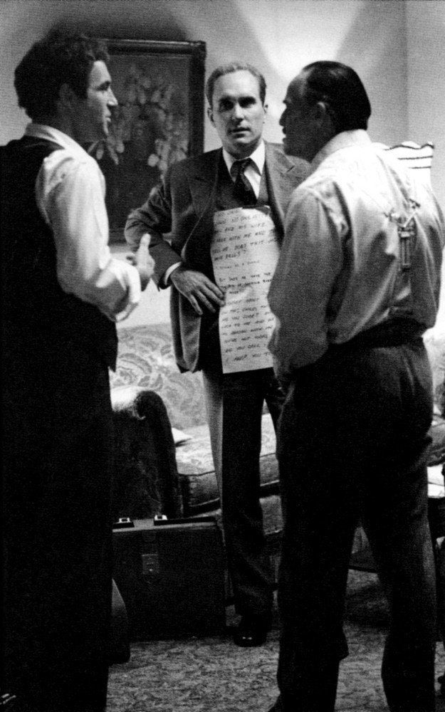 Three Men In Formal Attire Stand In A Room, Engaged In Conversation, While One Holds A Large Sheet Of Paper With Handwritten Notes. The Setting Includes A Couch And Wall Art In The Background, Subtly Reminiscent Of An Auto Draft Brainstorming Session.