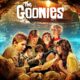 Goonies Cast Movie Poster - A group of children are gathered around a treasure map in an underground cave with the text "The Goonies" above them. An ocean and shipwreck can be seen in the background, bringing nostalgic echoes of the Goonies cast now.