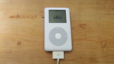 A white classic iPod with a circular click wheel rests on a wooden surface, connected via a cable, displaying the "Charging" screen along with the battery indicator icon.