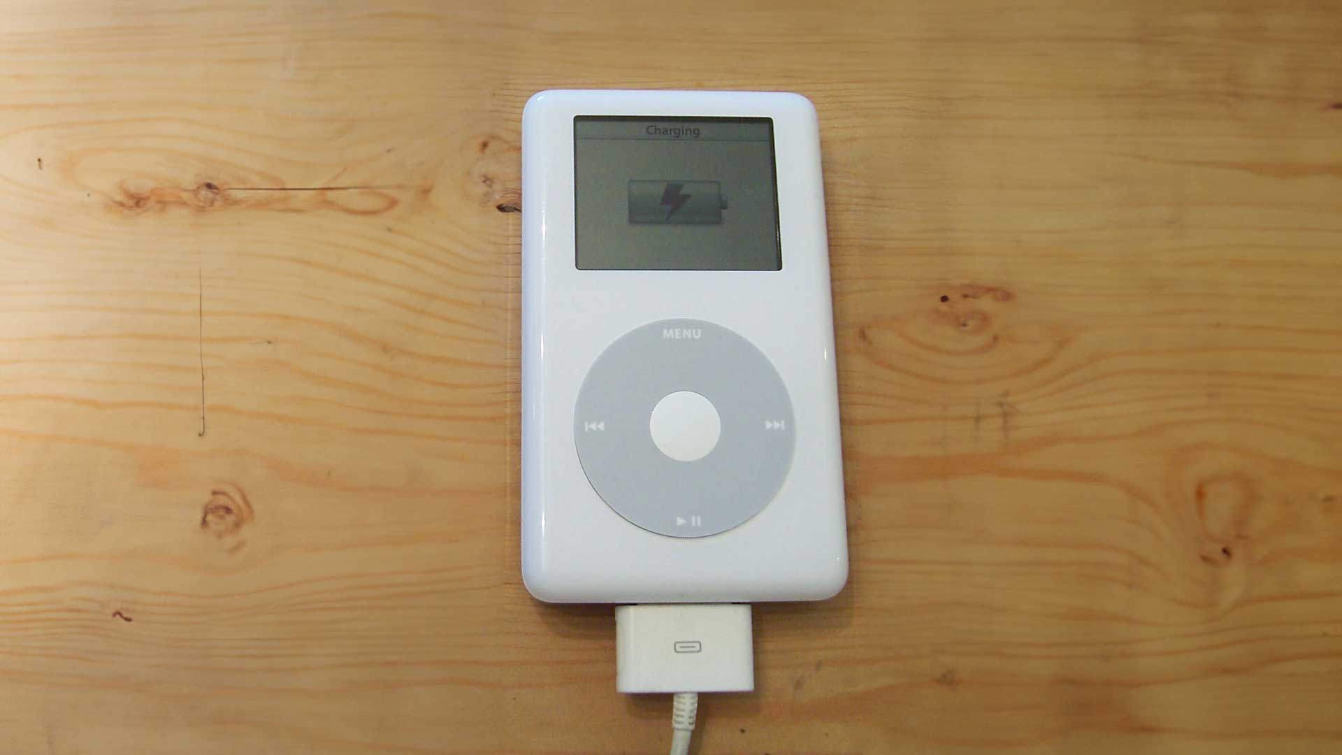 A White Classic Ipod With A Circular Click Wheel Rests On A Wooden Surface, Connected Via A Cable, Displaying The &Quot;Charging&Quot; Screen Along With The Battery Indicator Icon.