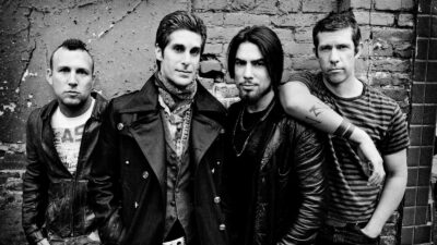 Jane's Addiction - Four men posing against a weathered brick wall; three are standing, one has his arm resting on another's shoulder, all dressed in casual and leather clothing. Black and white image capturing Jane's Addiction’s original lineup.