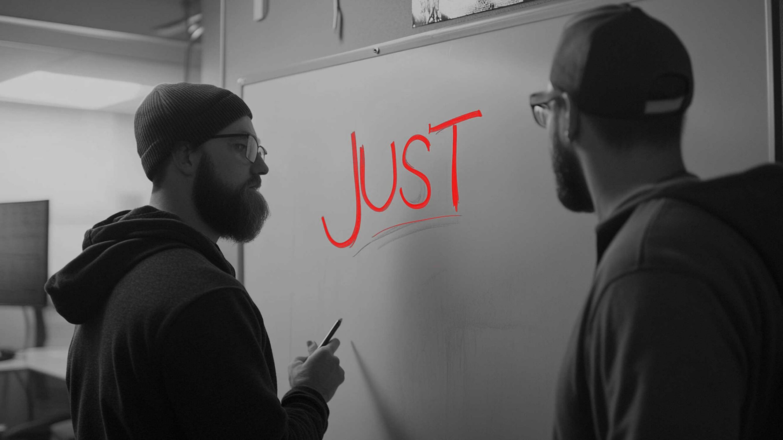 Two People In Hoodies And Caps Stand At A Whiteboard With The Word &Quot;Just&Quot; Written In Red, As If Plotting Their Next Move Like A Software Team Avoiding The Just Trap.