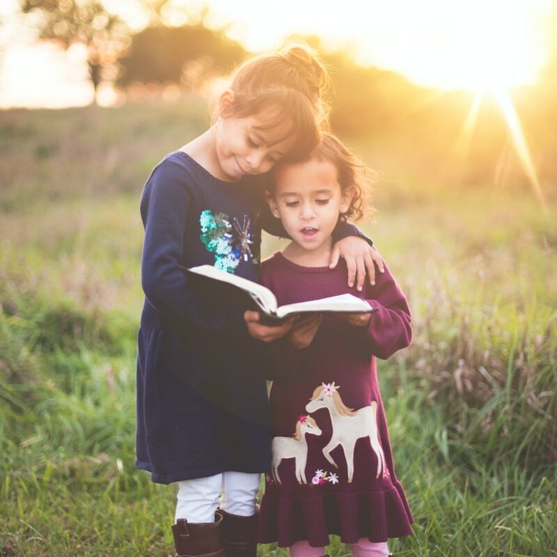 Happy Kids With A Book Or The Bible - Girl'S Left Hand Wrap Around Toddler While Reading Book During Golden Hour