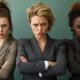 Three women in suits stand with arms crossed against a green background, each displaying a serious facial expression, embodying the strength of the Feminist Resurgence.