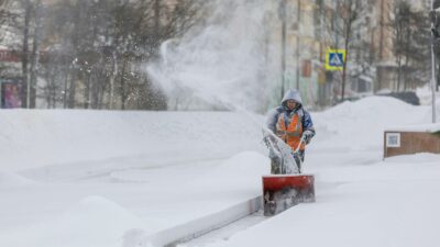 Person using a snow blower to clear a city sidewalk during a heavy winter snowfall.