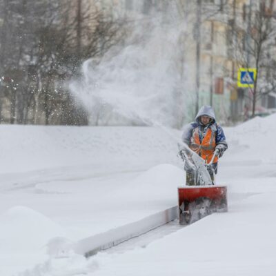 Person Using A Snow Blower To Clear A City Sidewalk During A Heavy Winter Snowfall.