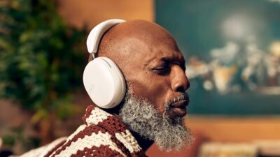 Sonos Ace Headphones Review - A bald man with a gray beard is wearing white headphones and appears to be enjoying music. Indoors with a blurry background featuring a painting and a green plant, he seems lost in the rich sound of his new Sonos Ace headphones.