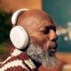 Sonos Ace Headphones Review - A bald man with a gray beard is wearing white headphones and appears to be enjoying music. Indoors with a blurry background featuring a painting and a green plant, he seems lost in the rich sound of his new Sonos Ace headphones.