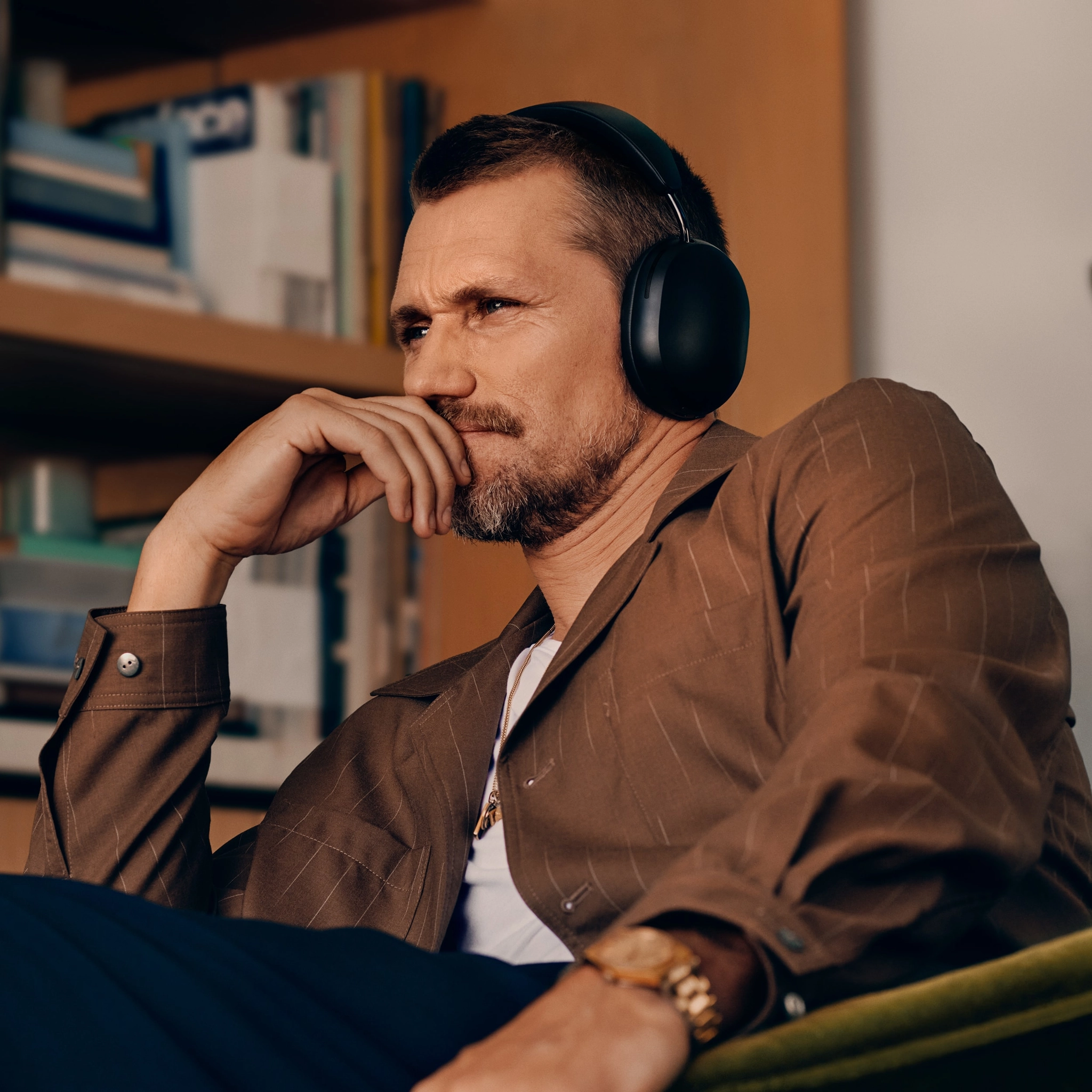 Sonos Ace Headphones - A Man With A Beard Is Wearing Large New Sonos Ace Headphones And A Brown Jacket While Sitting In A Chair, Appearing Thoughtful. There Are Bookshelves In The Background.