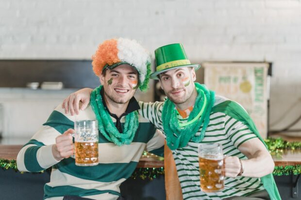 Happy Young Male Friends In Traditional Green And Yellow Outfits And Hats Cuddling And Smiling While Drinking Cold Beer While Listening To Celebration Songs For St. Patrick'S Day In A Pub.