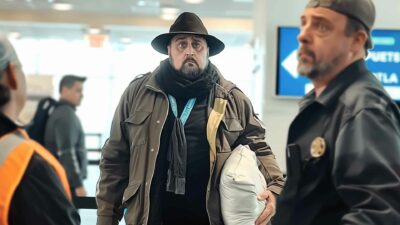 Airport Pillowcase Travel Hack Goes Wrong - A man wearing a black hat and brown jacket, employing a clever packing trick with a pillowcase, stands in an airport terminal alongside two other individuals.