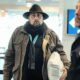 Airport Pillowcase Travel Hack Goes Wrong - A man wearing a black hat and brown jacket, employing a clever packing trick with a pillowcase, stands in an airport terminal alongside two other individuals.