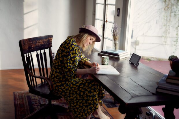 Woman In Yellow And Black Floral Dress Sitting On Brown Wooden Chair And Writing A Poem